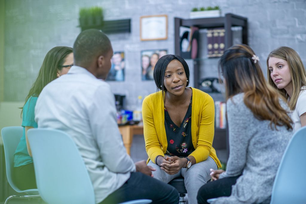 A diverse group of adults sit in a circle in their chairs and listen to and sympathize with each other with serious expressions.